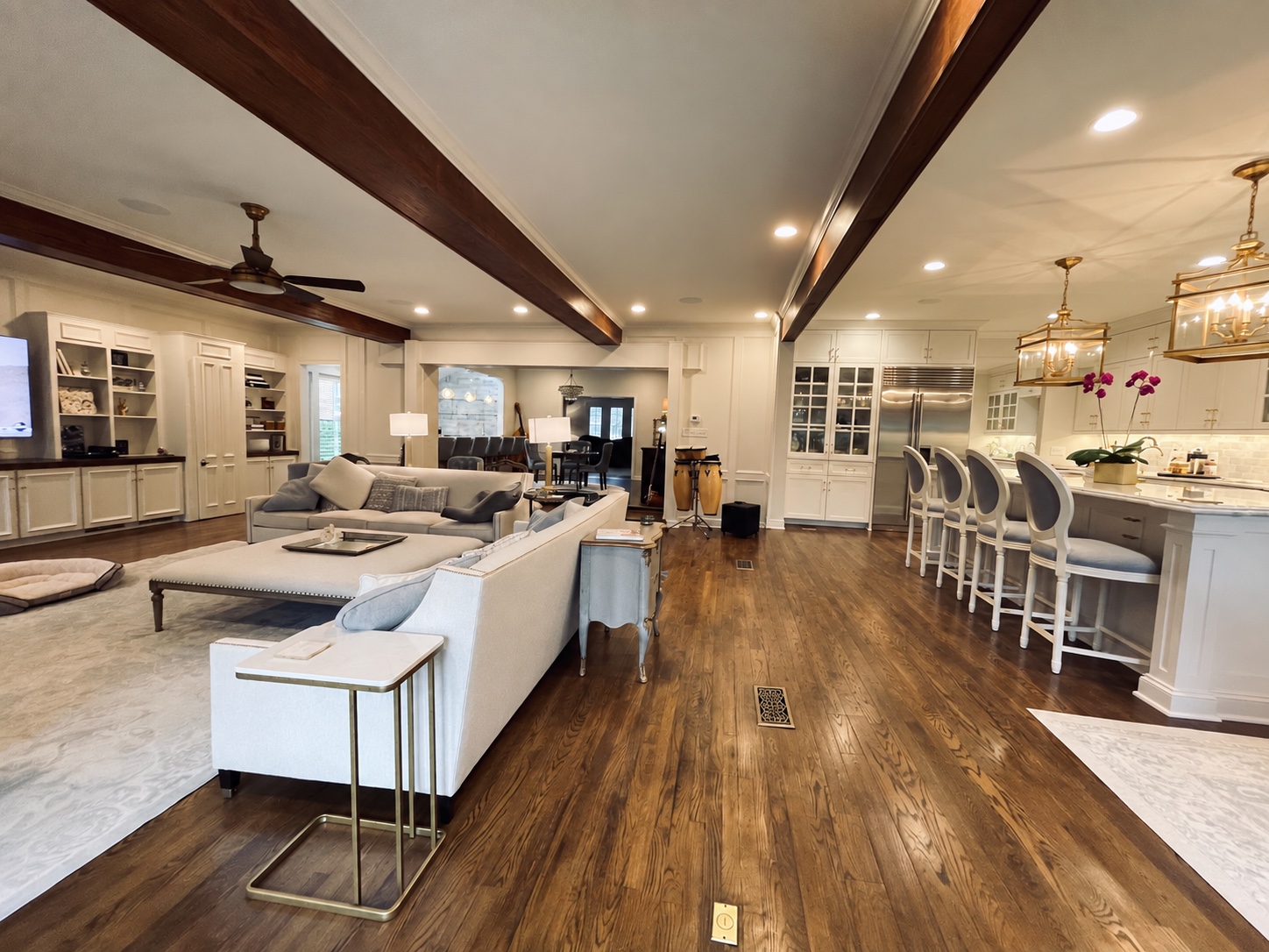 Furnished open-plan living room beside a white kitchen, with continuous wood flooring and dark ceiling beams