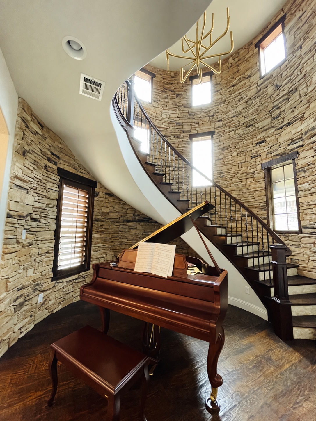 Curved staircase, grand piano, and gold chandelier in a stone-lined entry