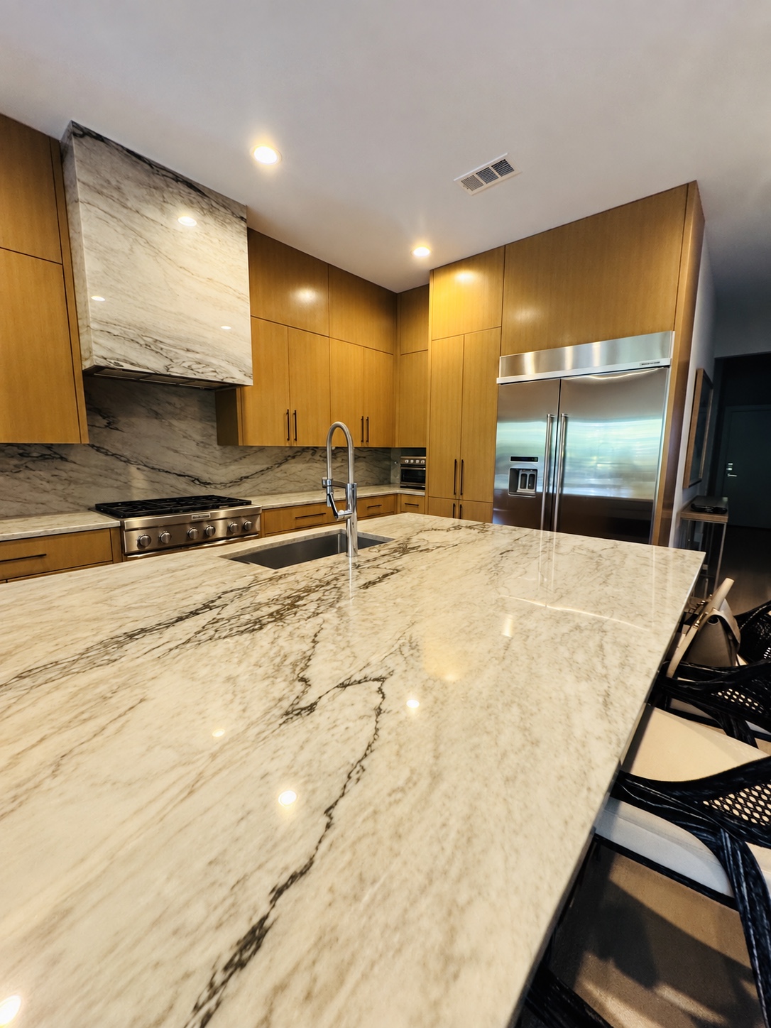 Kitchen with warm wood cabinetry, marble hood, clear marble island and stainless refrigerator