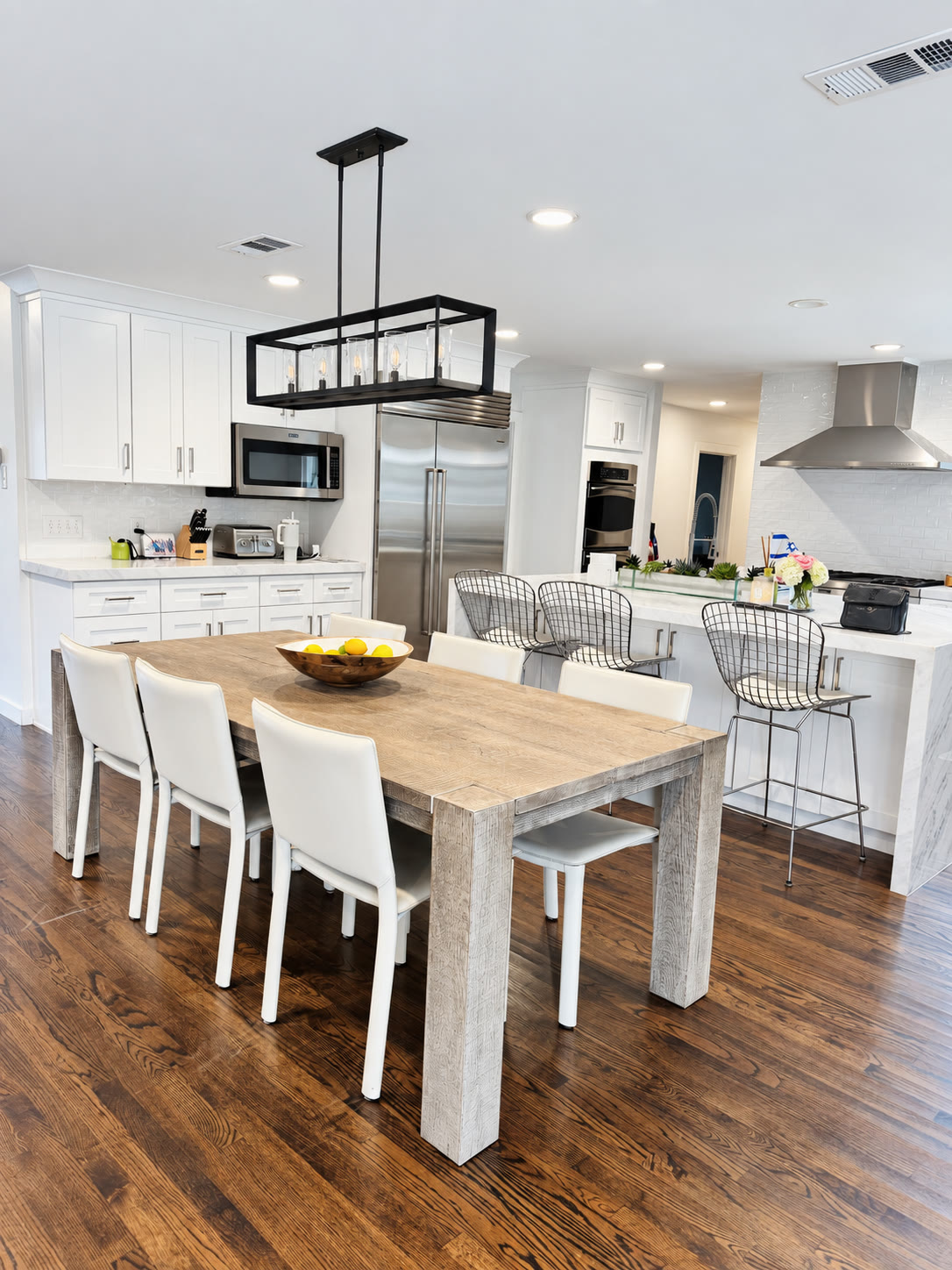 White kitchen and dining area with a black pendant, pale wood table and stainless appliances