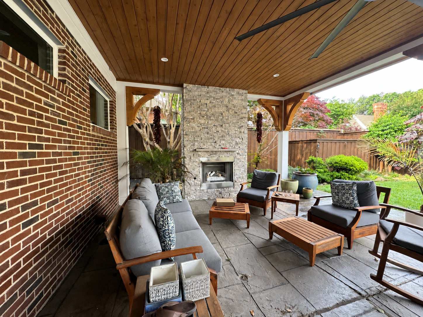Covered patio with wood ceiling, stone fireplace, gray lounge seating and a clear wooden bench