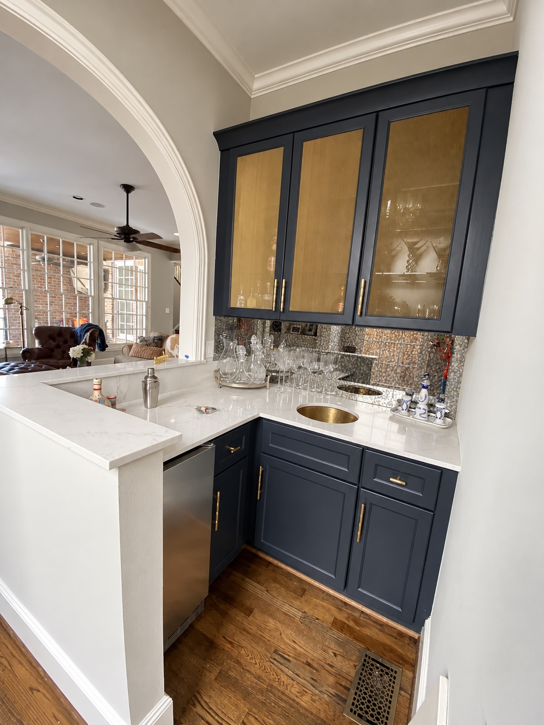 Home bar with charcoal-blue cabinetry, brass details, reflective tile backsplash and white counter