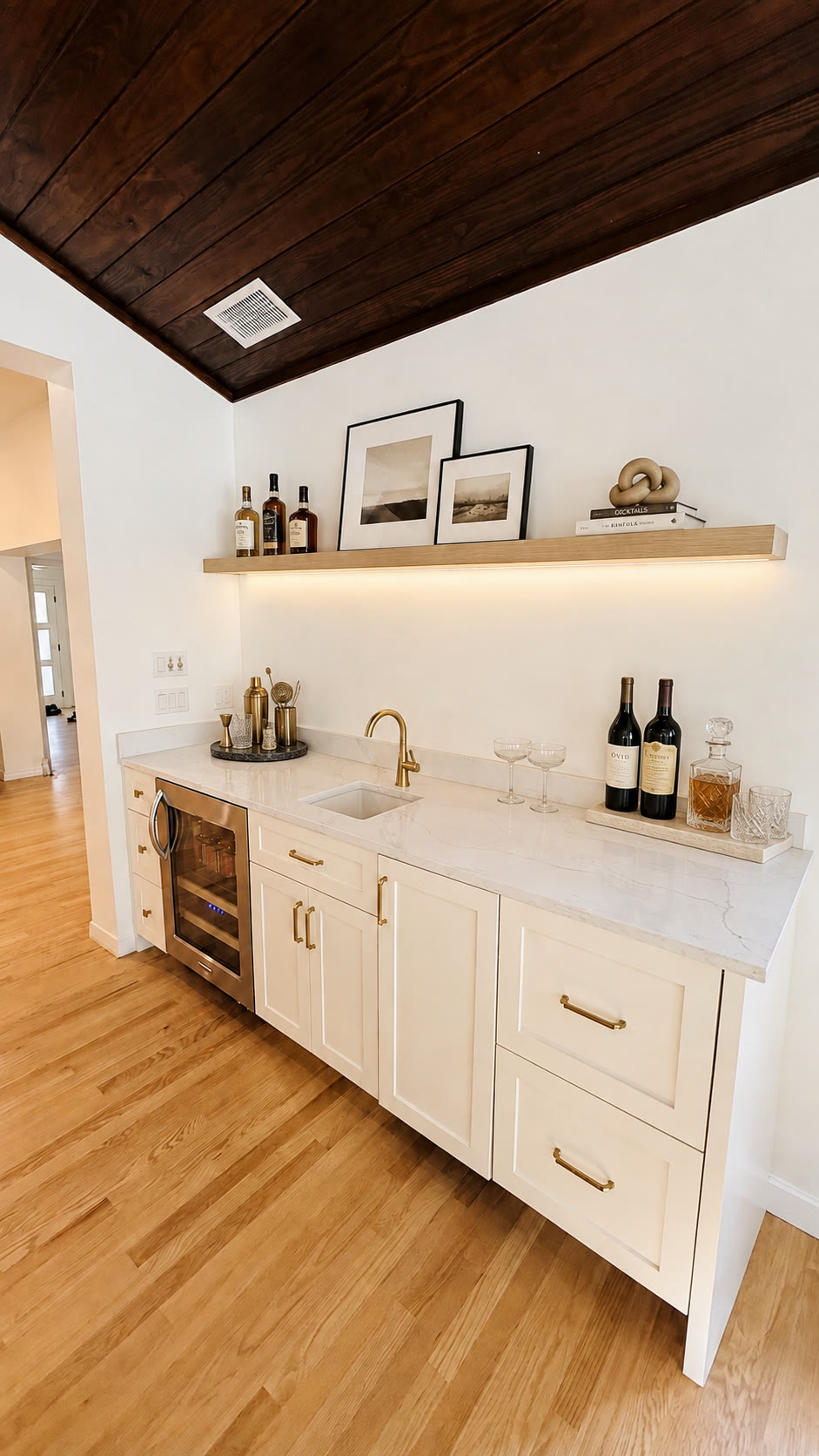 Digitally styled white wet bar with beverage refrigerator, wood ceiling, wine bottles and cocktail glassware