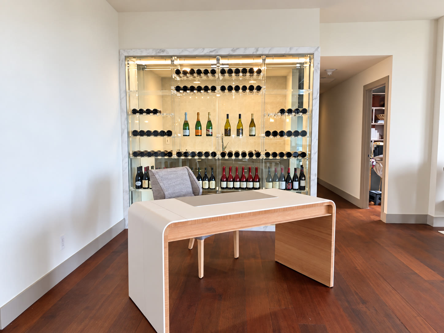 Illuminated glass wine display with bottle storage behind a modern white desk